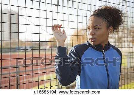 Picture - Young woman in sports jacket looking away through net at park. Fotosearch