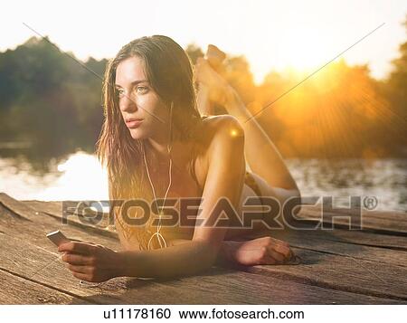 Stock Image - Young woman lying on jetty listening to mp3 player. Fotosearch