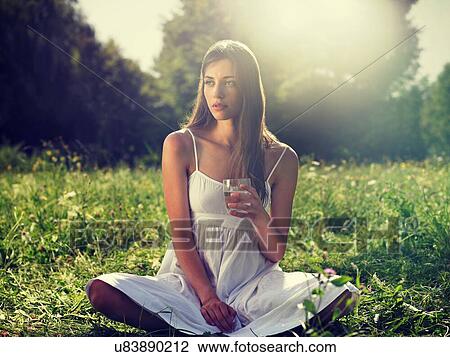 Stock Image - Young woman sitting in meadow cross-legged, holding drinking glass. Fotosearch