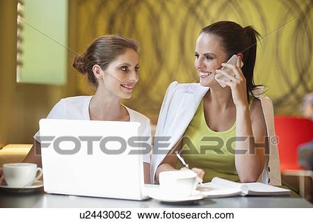 Young woman sitting with friend using cell phone at cafe View Large Photo Image Stock Image - Young woman sitting with friend using cell phone at cafe. Fotosearch