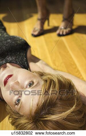 Stock Image - Adult woman lying on wood floor looking at viewer with feet of another woman in background wearing heels.. Fotosearch