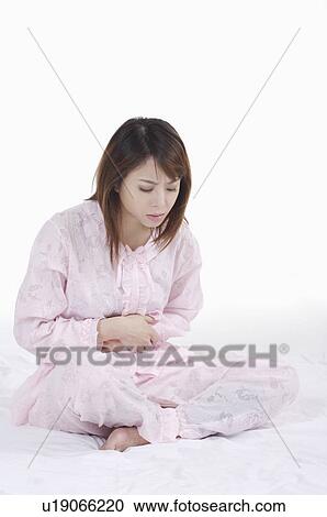 Stock Image - Young woman sitting on bed with hands on stomach. Fotosearch