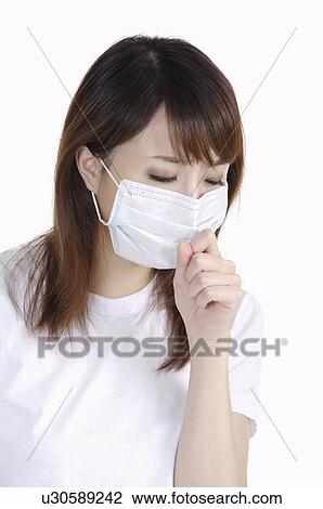 Stock Image - Young woman sneezing and wearing surgical mask. Fotosearch
