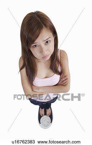 Stock Image - Young woman standing on weight scale and puckering of anxiety. Fotosearch
