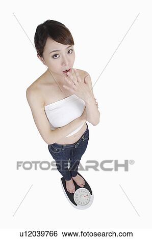 Stock Photograph - Young woman standing on weight scale and shocking with mouth open. Fotosearch