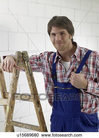 Stock Image - Man wearing work wear is standing next to a ladder. Fotosearch