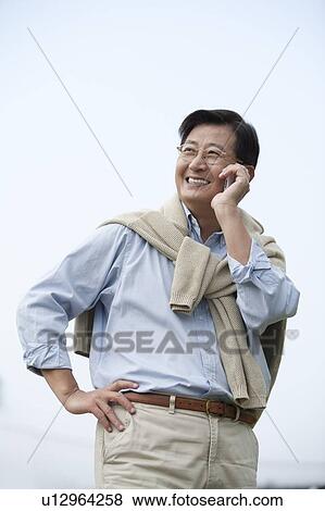 Stock Photo - Husband, Man standing with arms akimbo and using mobile phone with smile. Fotosearch