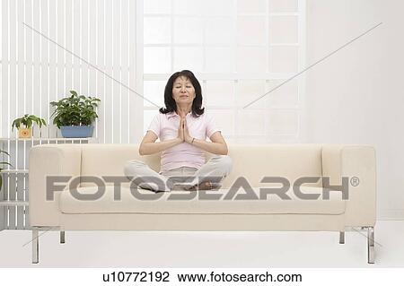 Stock Image - Wife, Woman making lotus position on sofa with eyes closed. Fotosearch