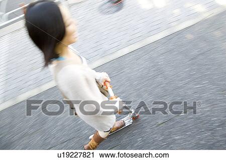 Stock Image - Young woman riding a kick scooter. Fotosearch