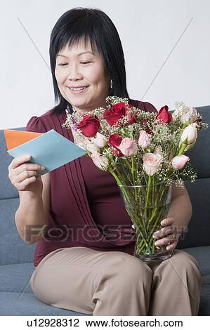Stock Image - Mature woman holding a flower vase and reading a greeting card. Fotosearch