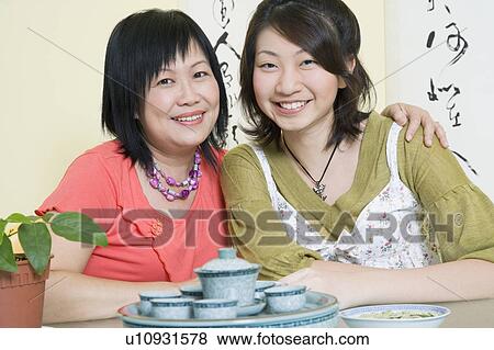 Stock Photo - Portrait of a mature woman sitting with her arm around her daughter at a table and smiling. Fotosearch
