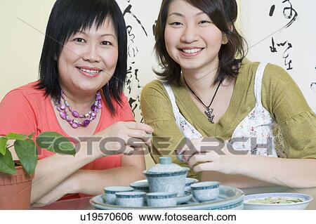 Stock Photograph - Portrait of a mature woman sitting with her daughter at a table and holding tea cups. Fotosearch