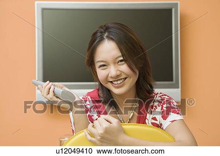 Stock Image - Portrait of a young woman holding a remote control and smiling. Fotosearch