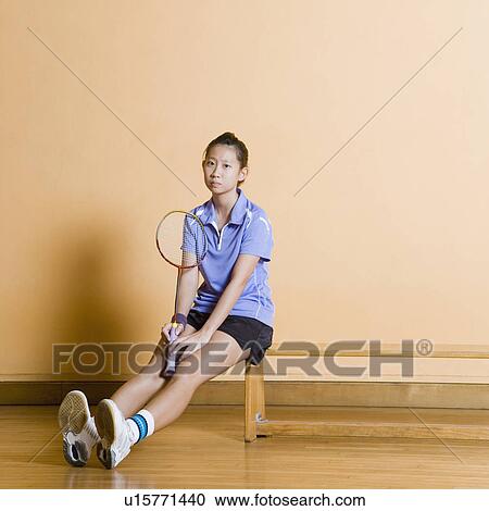 Stock Image - Portrait of a young woman sitting on a bench and holding a badminton racket. Fotosearch