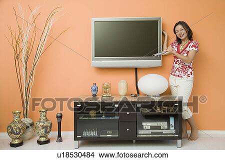 Young woman holding a remote control and standing near a cabinet View Large Photo Image Picture - Young woman holding a remote control and standing near a cabinet. Fotosearch