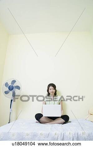 Picture - Young woman sitting cross-legged on the bed and using a laptop. Fotosearch