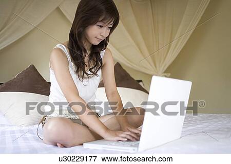 Stock Photo - Young woman sitting on a bed using laptop, Saipan. Fotosearch