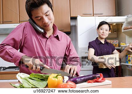 Stock Photo - Close-up of a mid adult man cutting vegetables while talking on a cordless phone and a young woman looking at him. Fotosearch