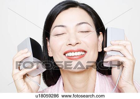 Stock Photograph - Close-up of a mid adult woman holding speakers upto her ear. Fotosearch