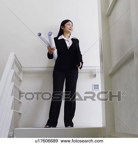 Stock Photo - Low angle view of a female architect standing on the staircase and holding rolled up blueprints. Fotosearch