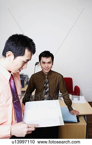 Stock Image - Portrait of a businessman smiling with another businessman reading a file. Fotosearch