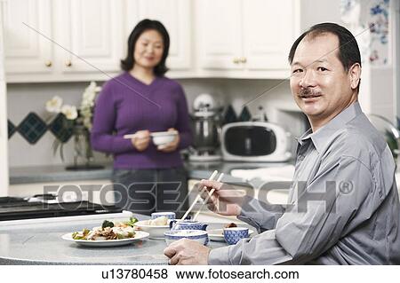 Stock Photo - Portrait of a senior man holding a pair of chopsticks with a mature woman standing behind him. Fotosearch