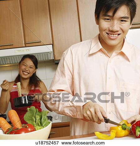 Stock Photo - Portrait of a young man cutting vegetables in the kitchen with a young woman holding noodles in the background. Fotosearch