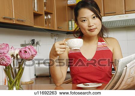 Stock Image - Portrait of a young woman holding a newspaper and a cup of tea at a kitchen counter. Fotosearch
