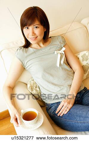 Stock Photo - Portrait of a young woman sitting on a couch and holding a cup of tea. Fotosearch