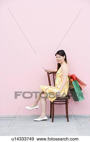 Stock Photo - Side profile of a young woman sitting on a chair and holding a mobile phone. Fotosearch
