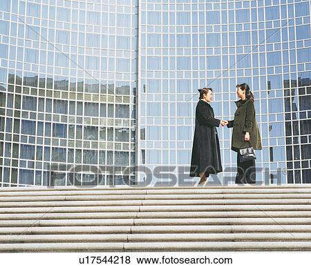 Stock Photo - Side profile of two young women standing and shaking hands in front of a building. Fotosearch