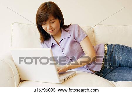 Stock Image - Young woman lying on a couch and working on a laptop. Fotosearch