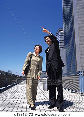Stock Image - Businessman and businesswoman pointing and looking at something by the high rise building, Low Angle View. Fotosearch