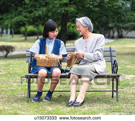 Stock Image - Grandmother and granddaughter sitting together on a bench, Front View. Fotosearch
