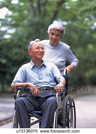 Stock Photography - Senior adult man on a wheelchair and his wife pushing it, Front View. Fotosearch
