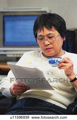 Stock Photo - Senior adult woman reading documents with a cup in hand, Front View. Fotosearch