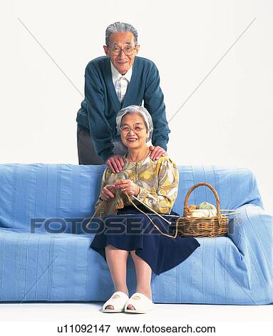 Stock Photo - Senior adult woman sitting on a sofa and her husband standing behind, Front View. Fotosearch