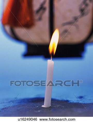 Stock Photograph - Burning candle and paper lantern, front view, blue background, differential focus. Fotosearch