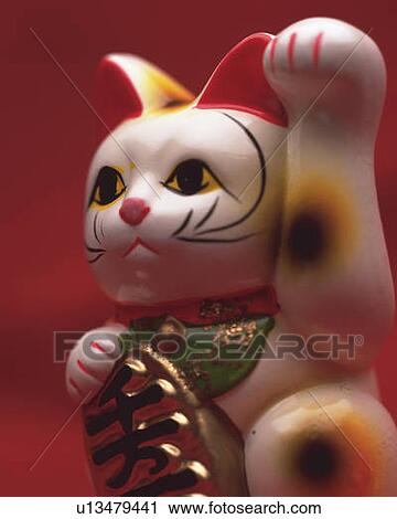 Stock Image - Doll of cat with a beckoning paw, side view, red background, differential focus. Fotosearch