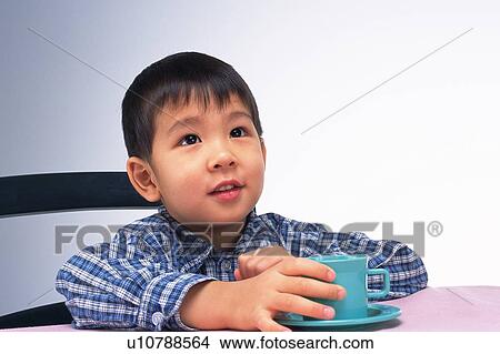 Picture -  a Little Boy Holding a Cup at the Table, Looking Up, Front View. Fotosearch