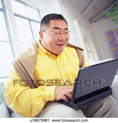 Stock Image -  a Mid Adult Man Using a Laptop, Sitting on a Sofa, Smiling, Low Angle View. Fotosearch