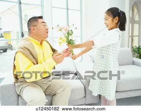 Stock Image - Daughter Giving a Bunch of Flowers to her Father who is Sitting on the sofa, Smiling, Side View. Fotosearch