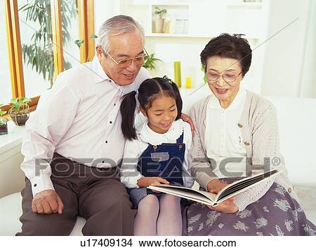 Picture - Grandparents and granddaughter Sitting on a Sofa in the living room Reading a Book, Smiling, Front View. Fotosearch