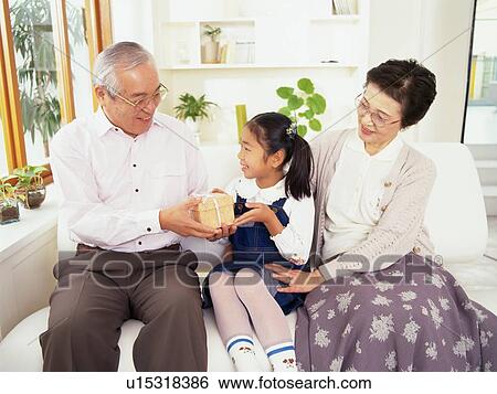 Stock Photograph - Grandparents and granddaughter Sitting on a Sofa in the living room, Grandfather passing a Gift to his Granddaughter, Smiling, Front View. Fotosearch