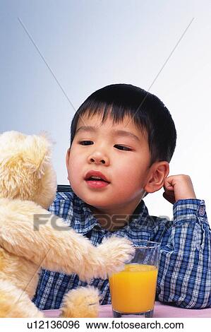 Stock Photograph - Little Boy playing with a teddy bear, While Drinking a Glass of Orange Juice, Front View. Fotosearch