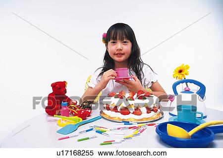 Stock Photography - Little Girl holding a saucer and a cup While sitting at a table with a birthday cake and some kitchenware, looking at camera, Smiling, Front View. Fotosearch