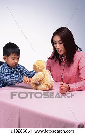 Stock Image - Mother and her Little son playing with a teddy bear on a Table, Side View. Fotosearch
