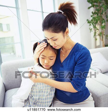 Stock Image - Mother holding daughter's head, Checking for Fever With a Thermometer, Side View, Differential Focus. Fotosearch