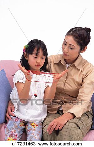 Stock Image - Mother looking at her daughter Who's playing cat's cradle, sitting on the sofa. Front View. Fotosearch