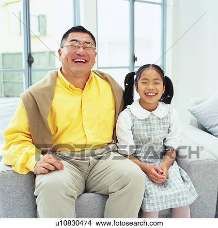 Picture - Portrait of a father and his daughter sitting on sofa in the Living Room, smiling, Looking at Camera, Front View. Fotosearch